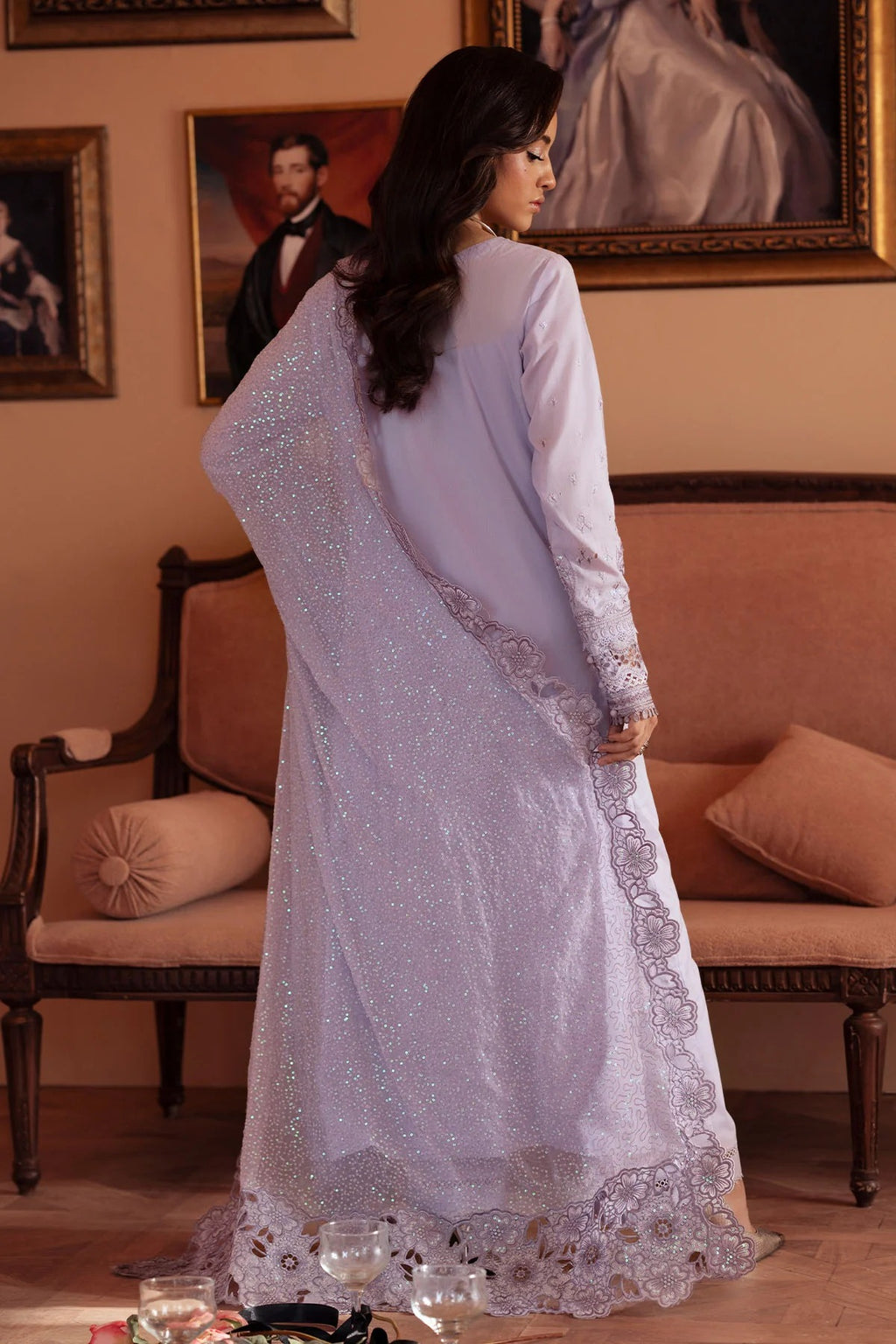 Woman in a white embroidered traditional outfit standing in a room with ornate furniture and portraits.