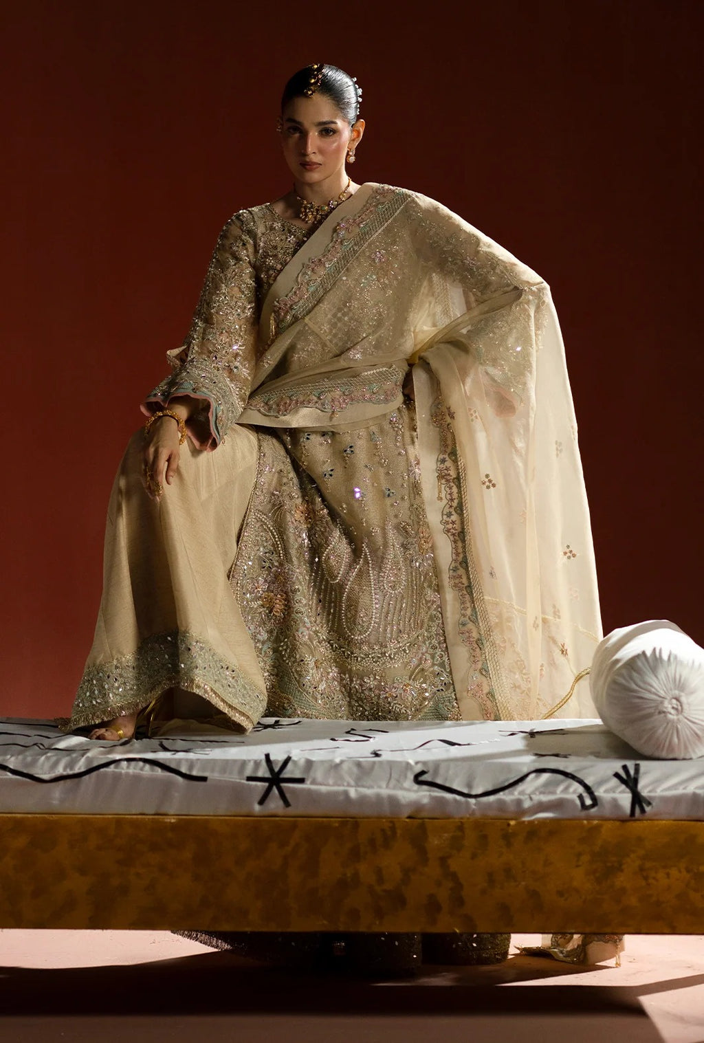 A model wearing a traditional beige and gold heavily embroidered ethnic outfit with a sheer draped shawl, seated on a decorated platform against a dark reddish-brown background.