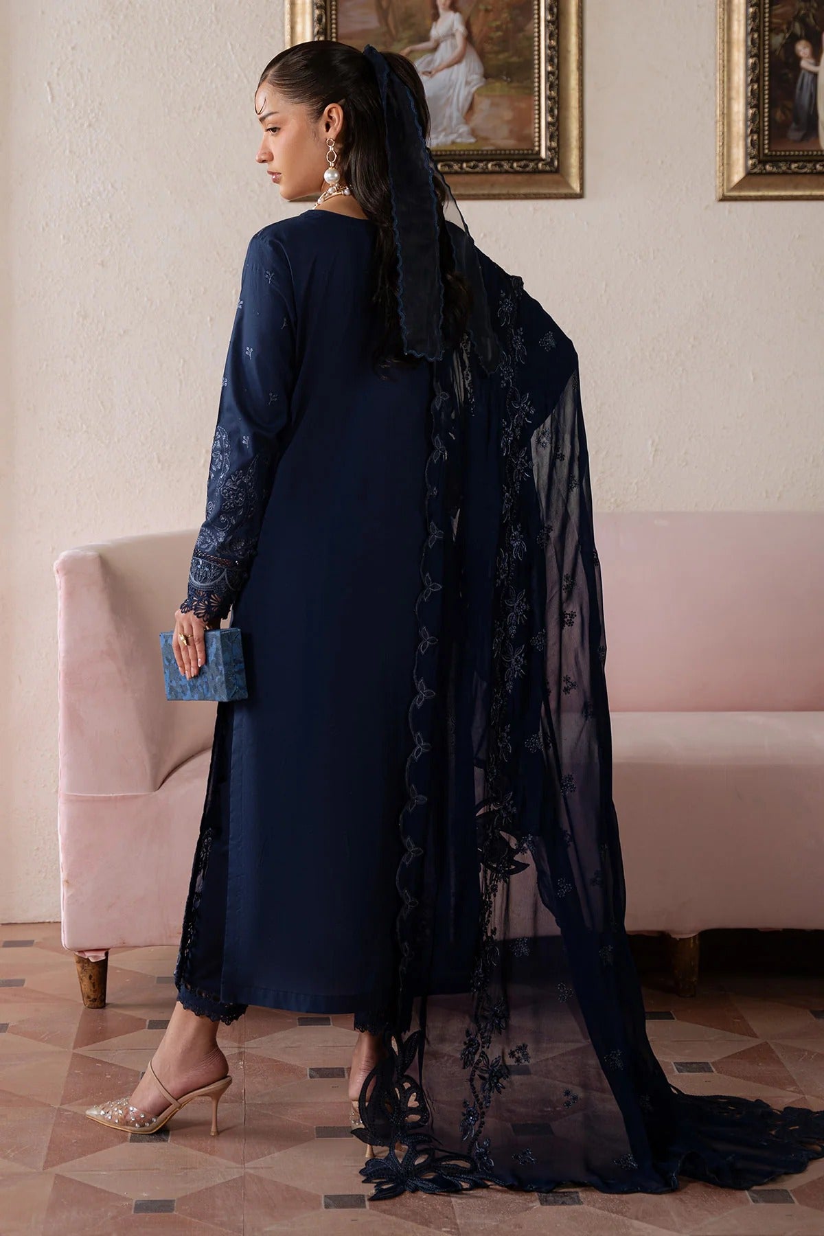Woman in a blue dress with a black shawl standing in a room with pink walls and framed pictures.