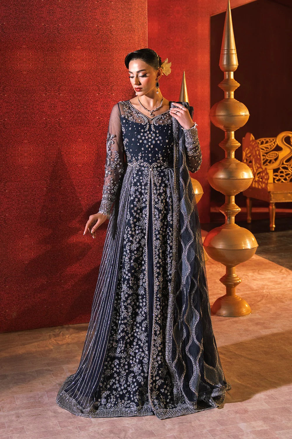 Woman in a dark blue embroidered dress standing in a room with red walls and gold decorative elements.