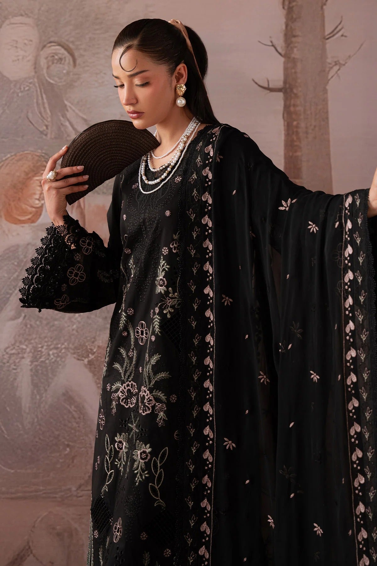 Woman in a black embroidered outfit holding a fan, standing against a decorative wall.