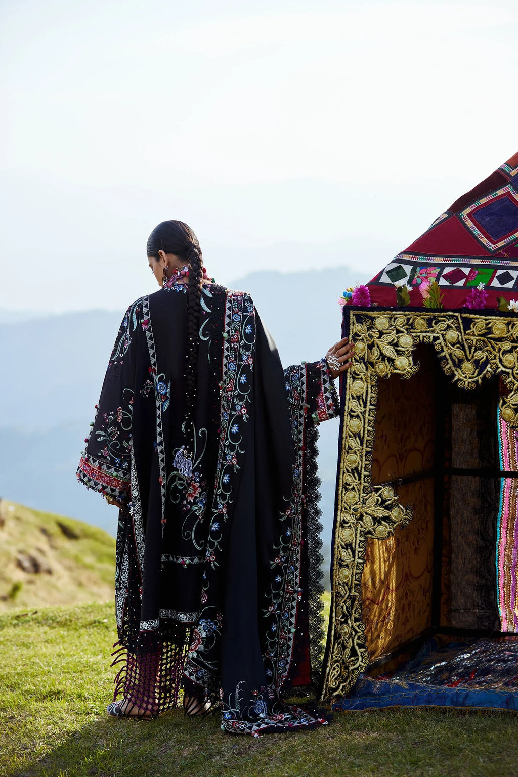 Person in a long, embroidered black and gold coat standing next to a colorful tent on a grassy hillside.