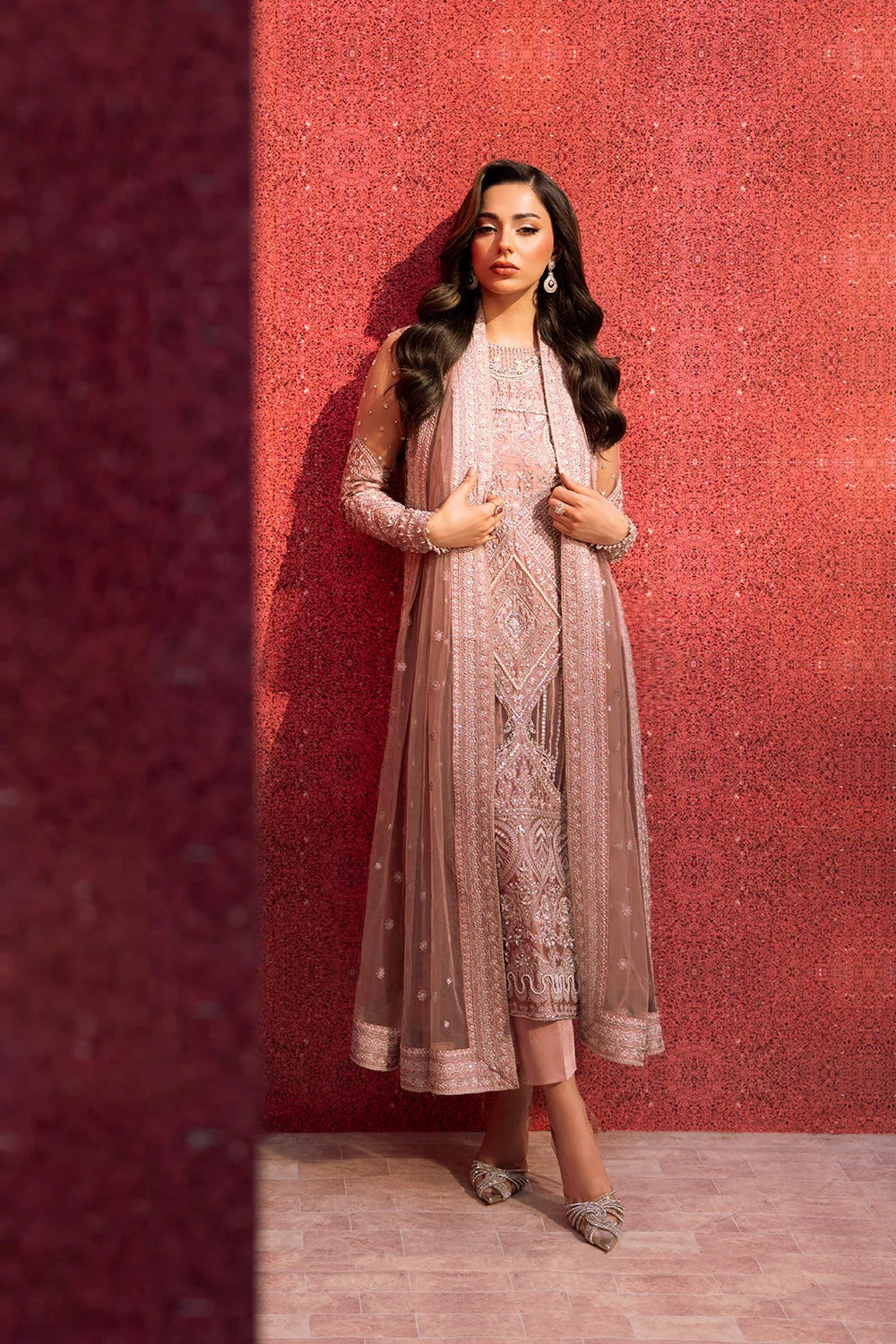 Woman in a pink embroidered outfit standing against a red patterned wall.
