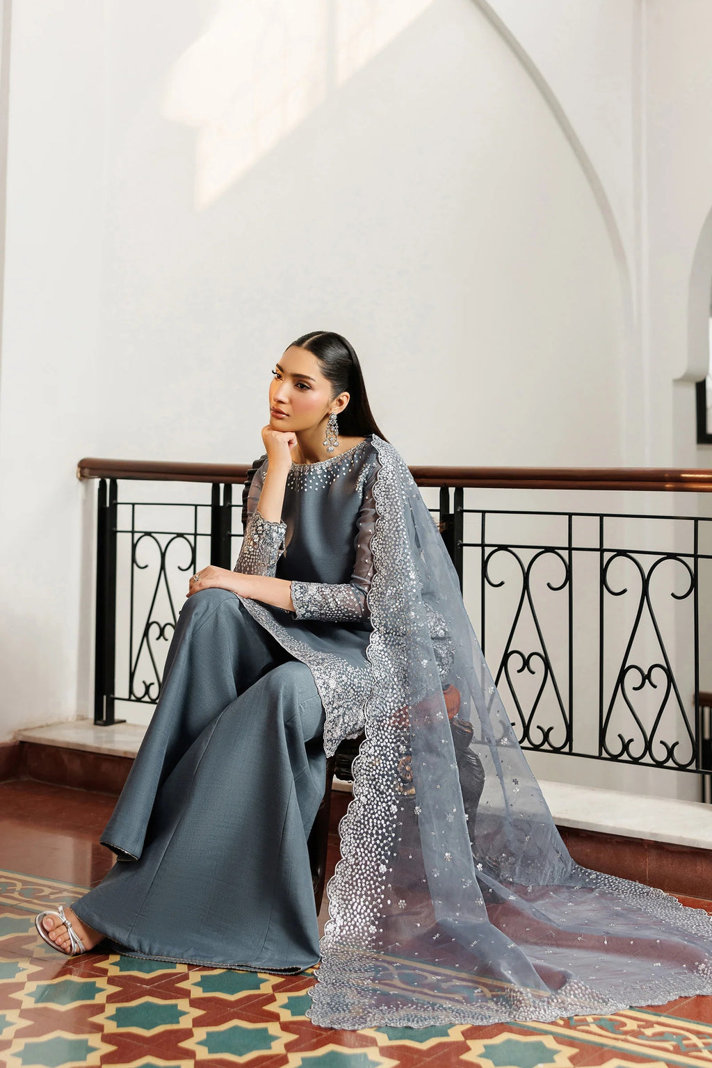 Woman in a gray traditional outfit with a long, embellished scarf sitting on a decorative floor.