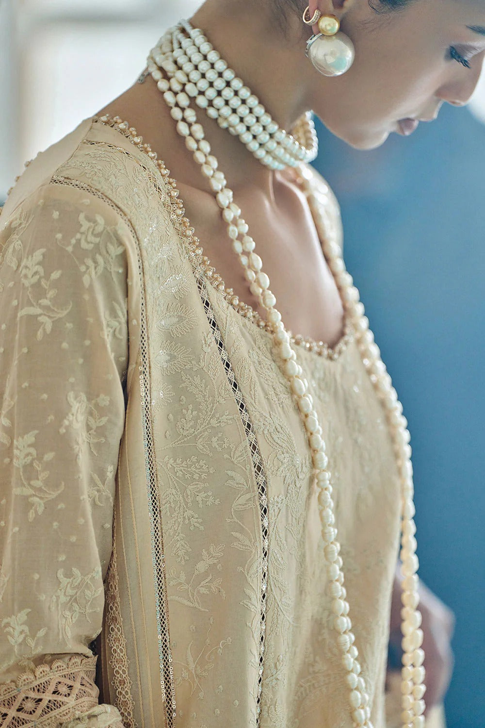 Woman wearing a beige embroidered outfit with pearl jewelry against a blurred background
