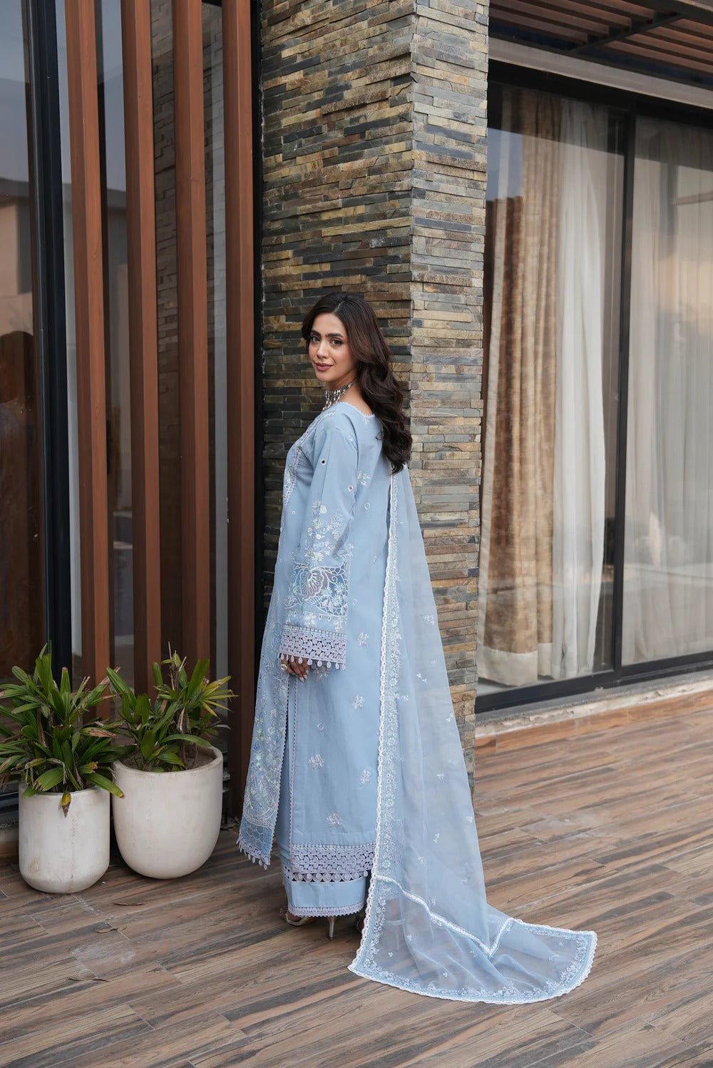 Woman in a light blue traditional outfit standing on a wooden deck with plants and a stone wall in the background.
