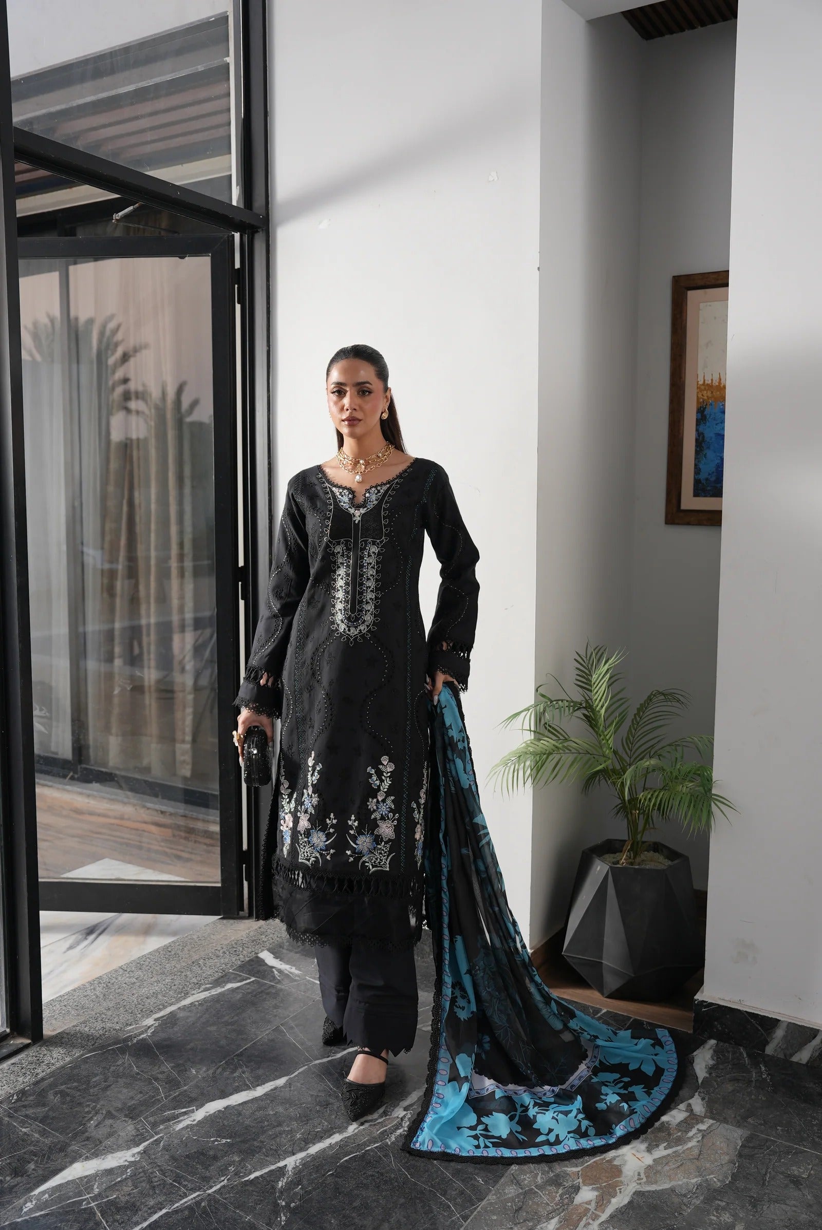 Woman in a black traditional outfit with a blue dupatta standing in a modern indoor setting.