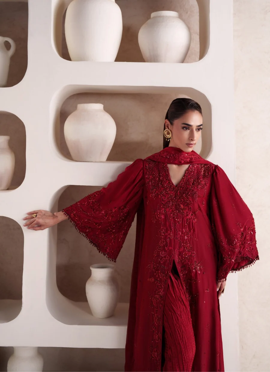 A woman models a deep red embroidered gown with wide sleeves and a matching scarf, standing in front of a beige textured wall with built-in white ceramic vases.