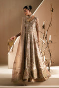 A woman models a traditional South Asian formal outfit consisting of a heavily embellished beige and gold anarkali-style gown with a matching sheer cape, styled with gold jewelry and a gold clutch bag against a warm beige studio background.