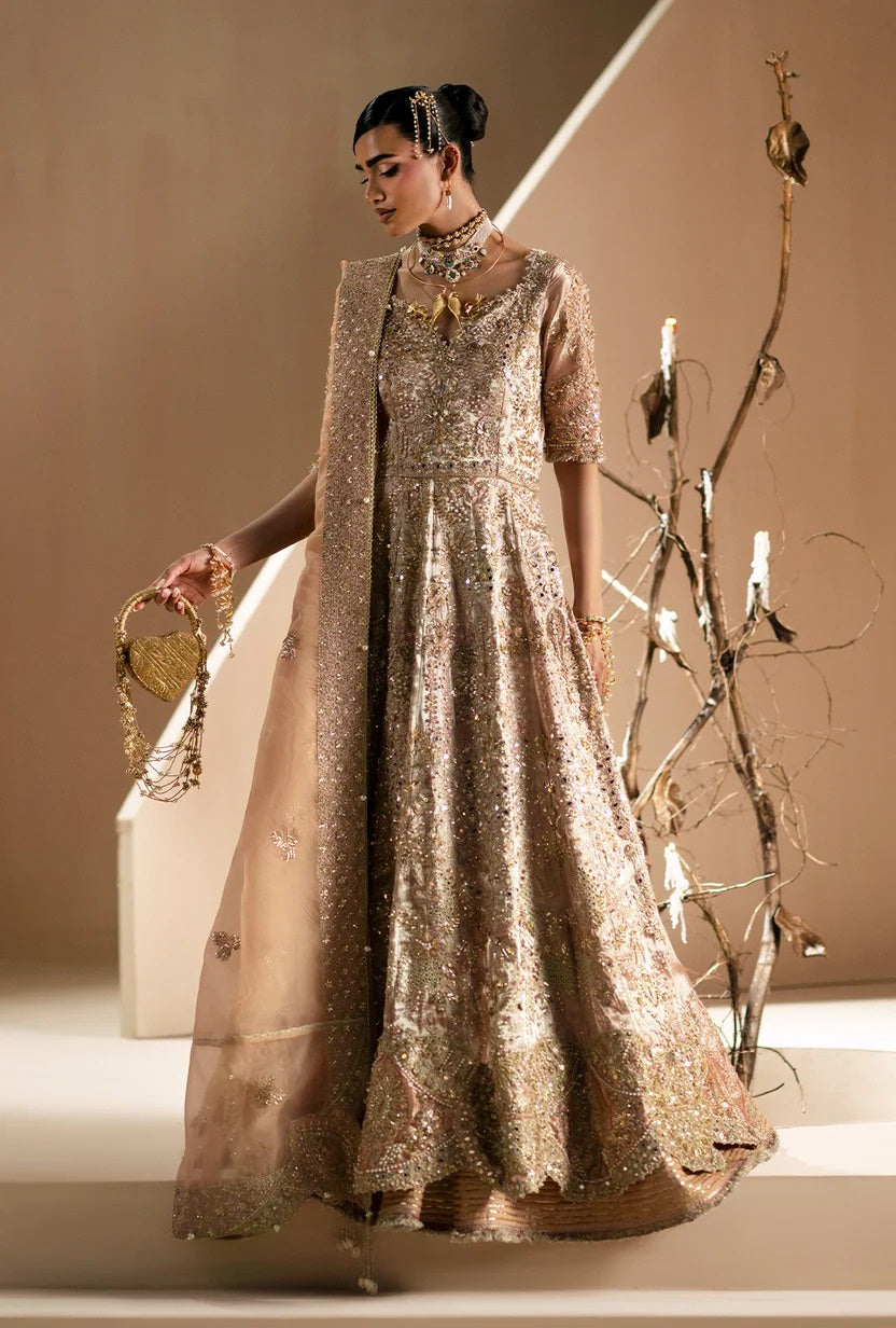 A woman models a traditional South Asian formal outfit consisting of a heavily embellished beige and gold anarkali-style gown with a matching sheer cape, styled with gold jewelry and a gold clutch bag against a warm beige studio background.