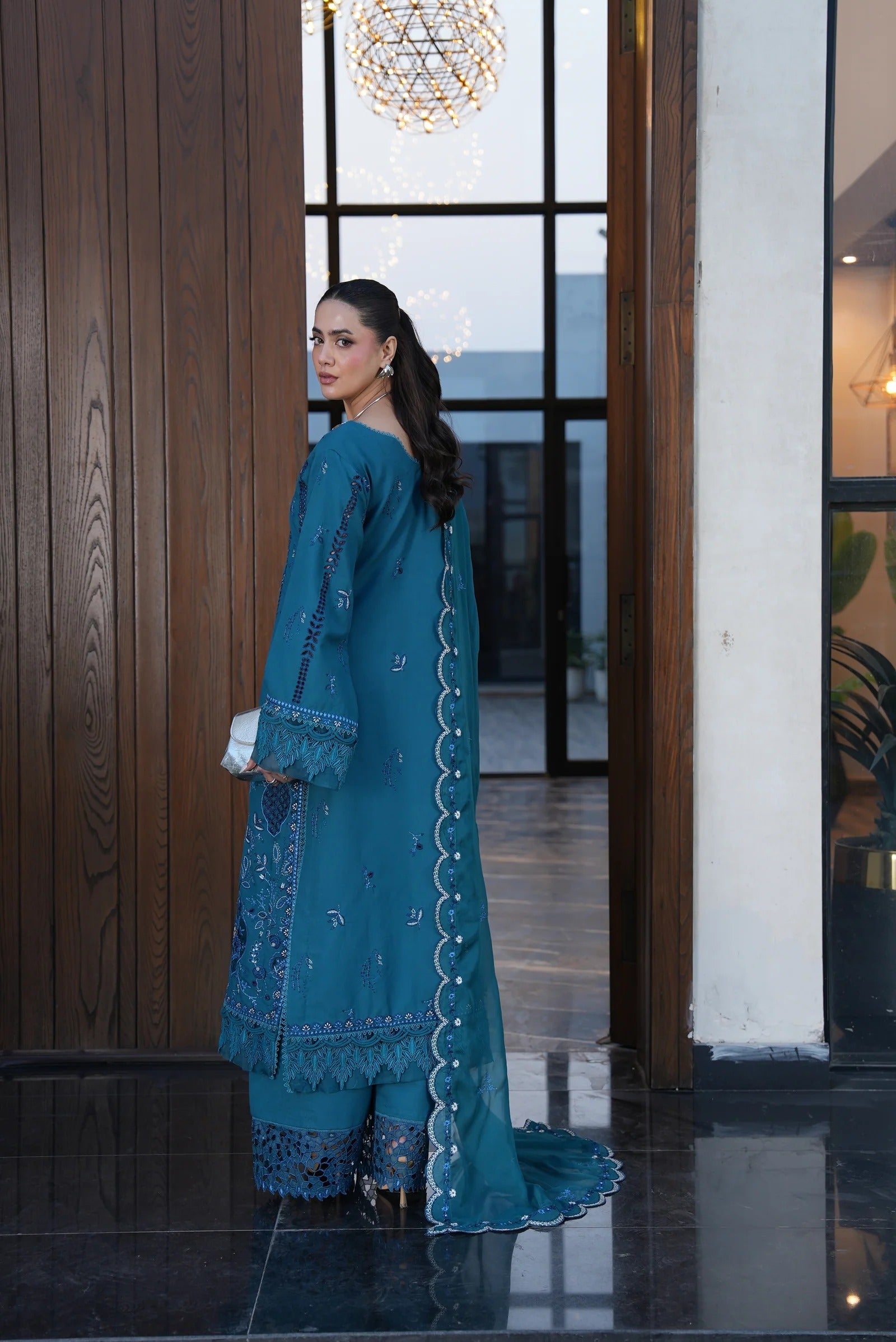 Woman in a teal traditional outfit standing in an elegant indoor setting.