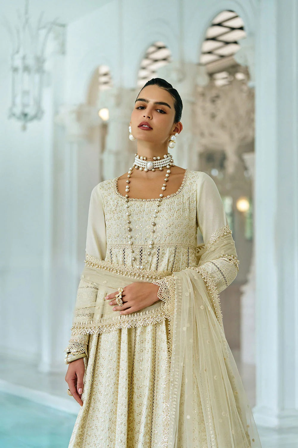 Woman in a cream traditional outfit with intricate patterns in an elegant indoor setting.