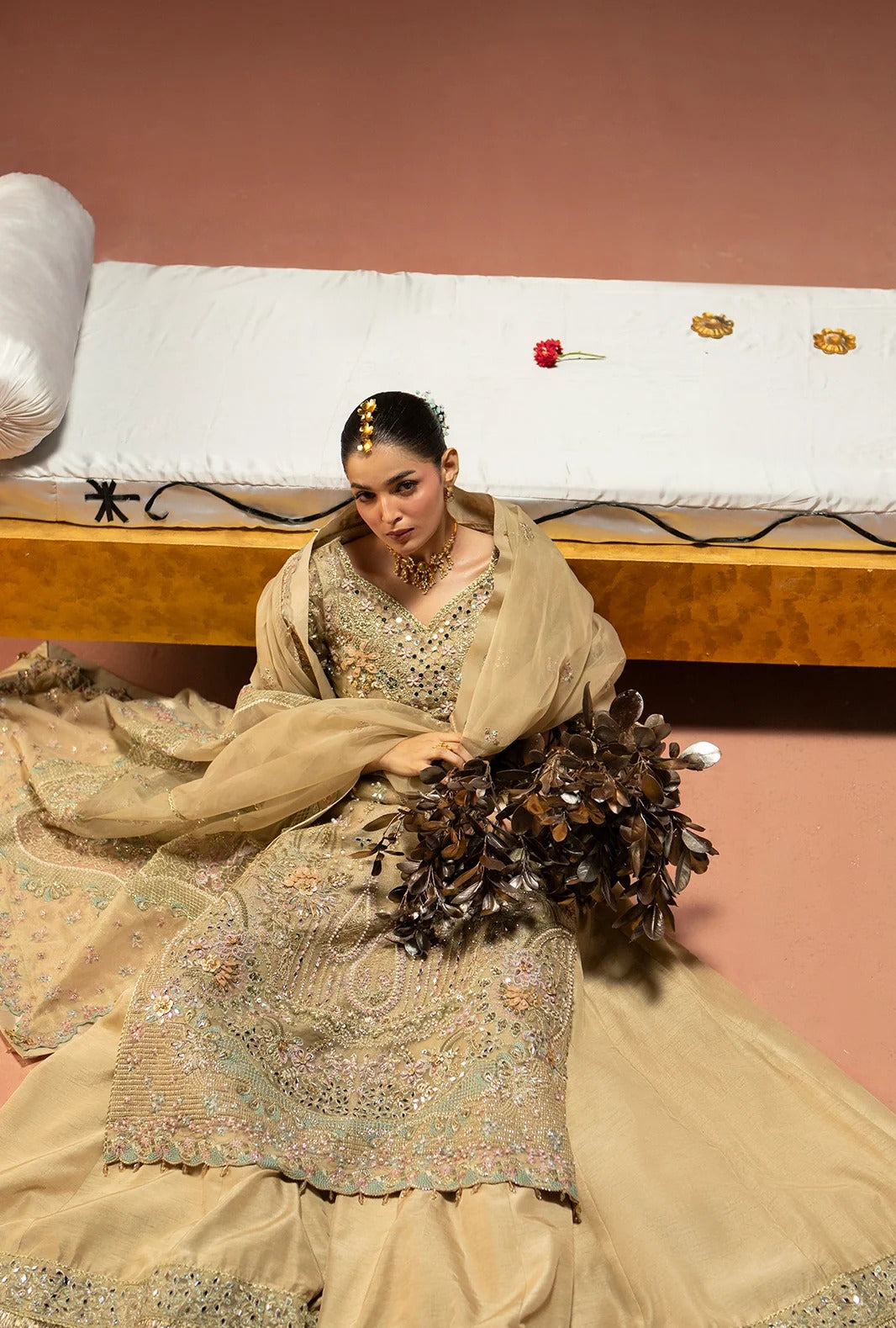 A woman seated on a bed wearing an ornate beige and gold embroidered traditional outfit with a sheer shawl, holding a dark floral bouquet in a bedroom setting.