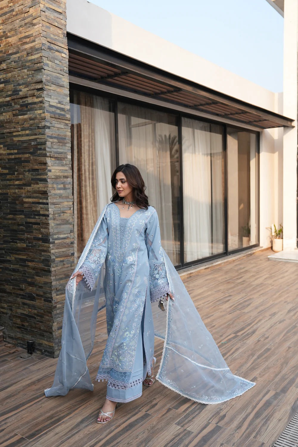 Woman in a light blue traditional outfit standing on a wooden deck with modern architectural background