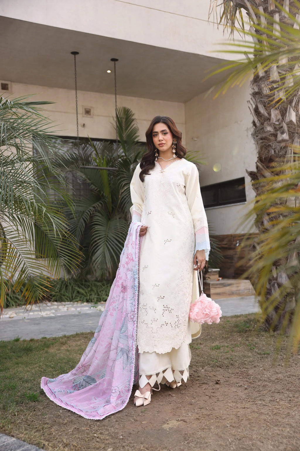 Woman in a white traditional outfit with a pink dupatta standing outdoors near a building and palm trees.