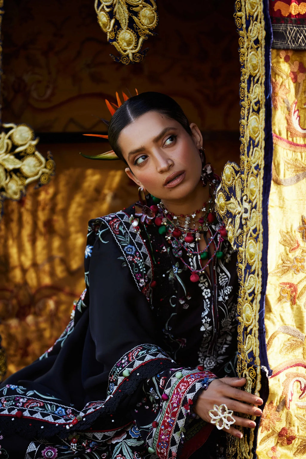 Woman in traditional attire with intricate patterns and jewelry, standing against a decorative background.