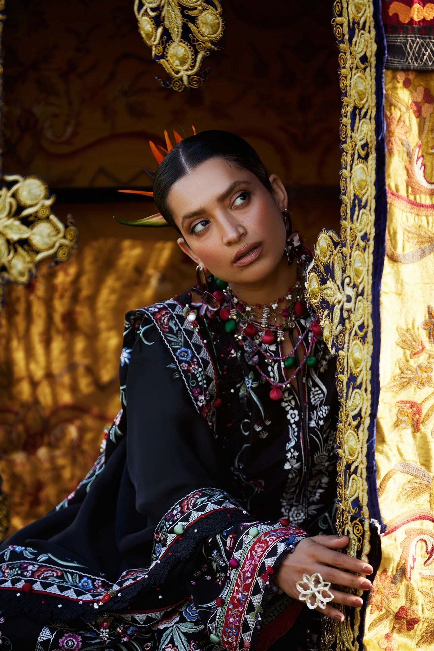 Woman in traditional attire with intricate patterns and jewelry, standing against a decorative background.