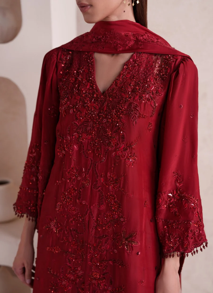 A close-up of a model wearing a deep red embroidered traditional dress with intricate floral embroidery and beadwork, featuring a draped collar and 3/4 sleeves.