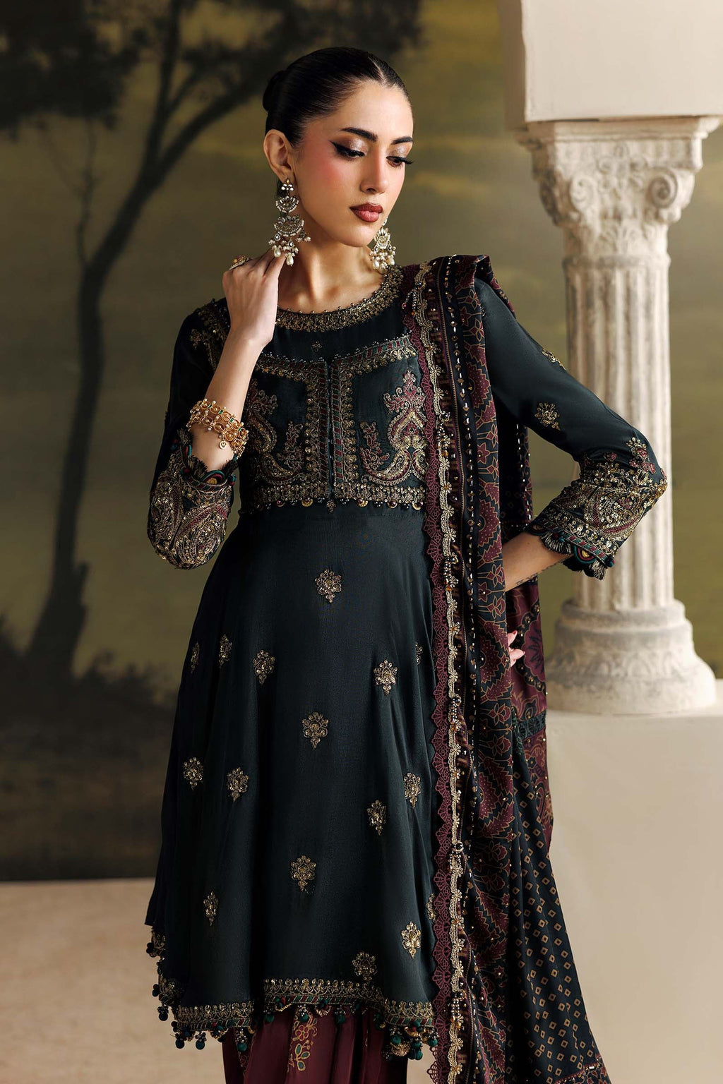 A woman models a dark charcoal green embroidered Anarkali kurta with a matching printed dupatta, styled with traditional jewelry in an elegant indoor setting with a classical column.
