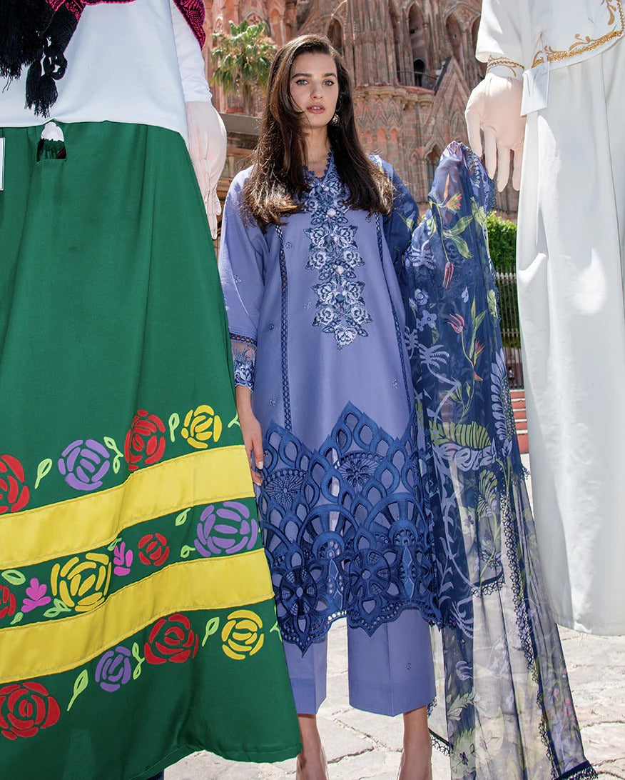 Woman in a blue embroidered dress with a colorful scarf, standing in front of a traditional building.