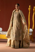 A woman models an ornate beige and gold traditional South Asian outfit with intricate embroidery, a matching sheer dupatta, and wide-leg pants against a warm brown studio backdrop with decorative gold furniture.