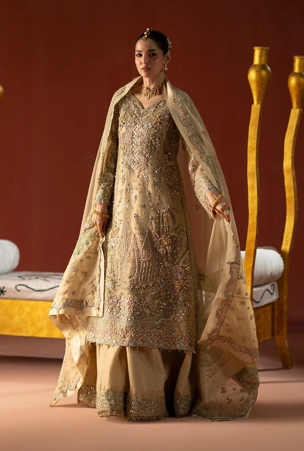 A woman models an ornate beige and gold traditional South Asian outfit with intricate embroidery, a matching sheer dupatta, and wide-leg pants against a warm brown studio backdrop with decorative gold furniture.