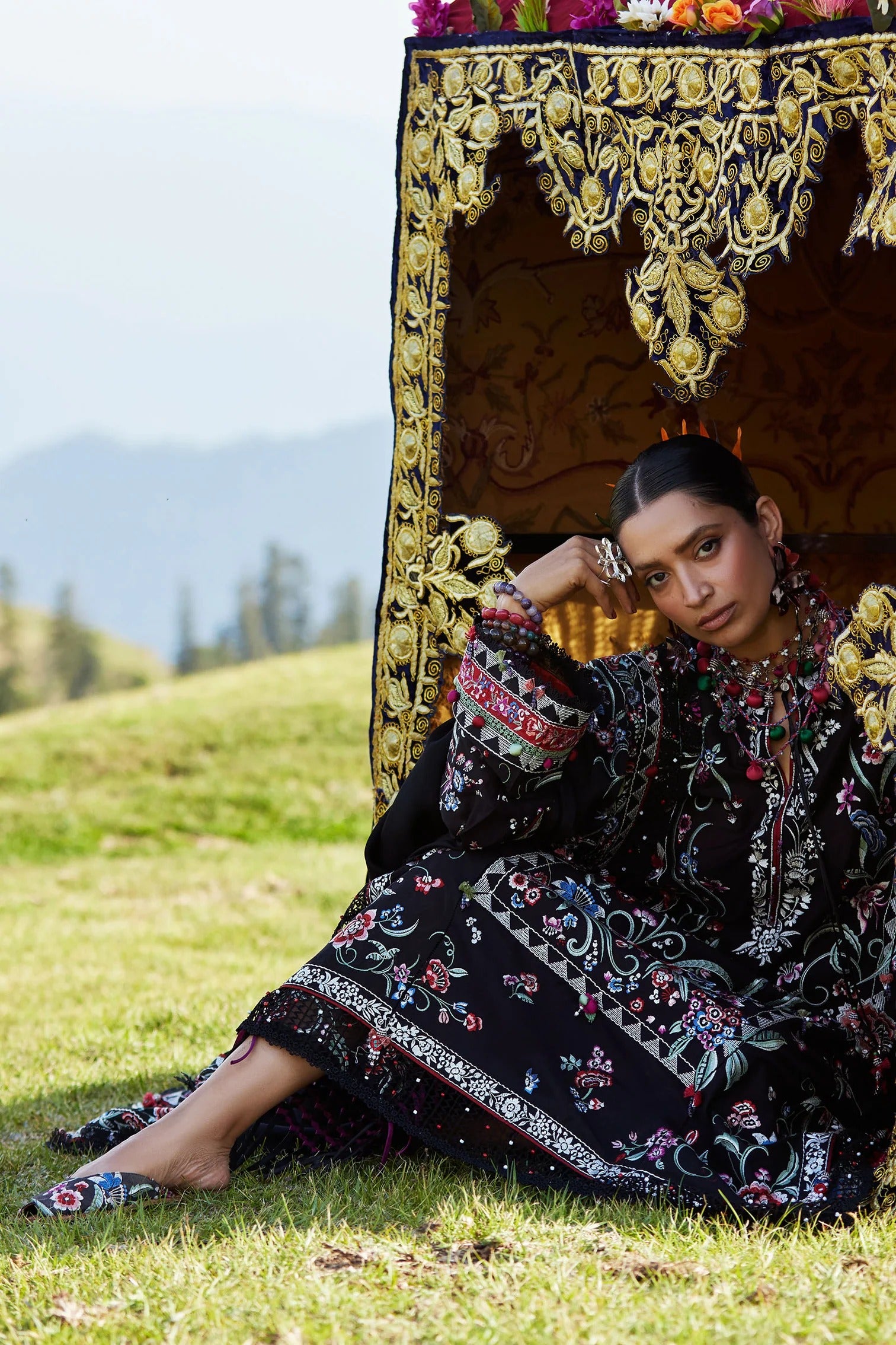Woman in traditional embroidered outfit sitting in front of a decorative screen with mountains in the background