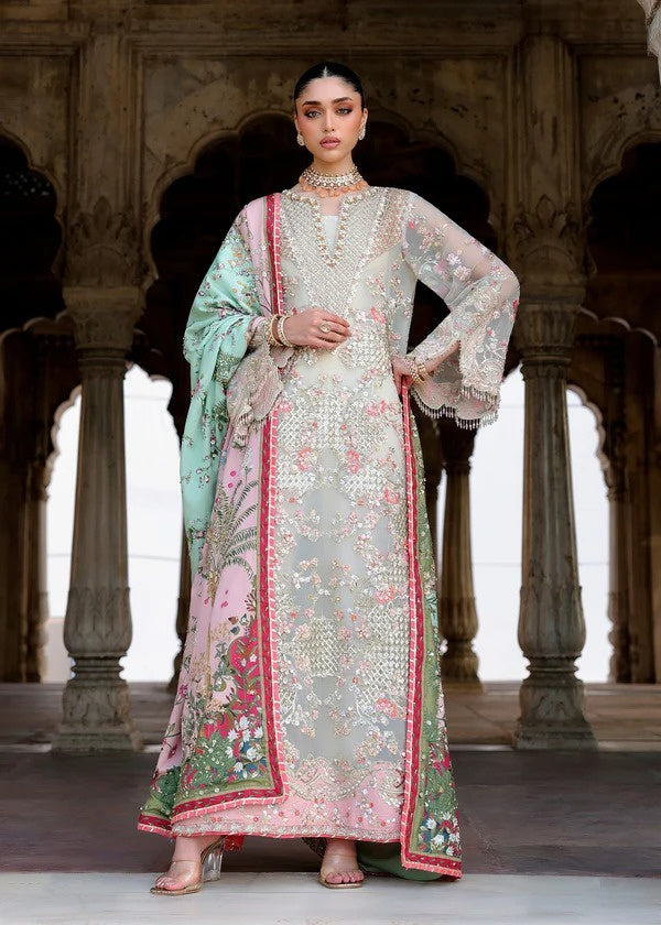 A woman models a traditional Indian salwar kameez suit with an embroidered light gray-green kurta, pink and green floral prints, and a mint green dupatta, set against a background of stone pillars.