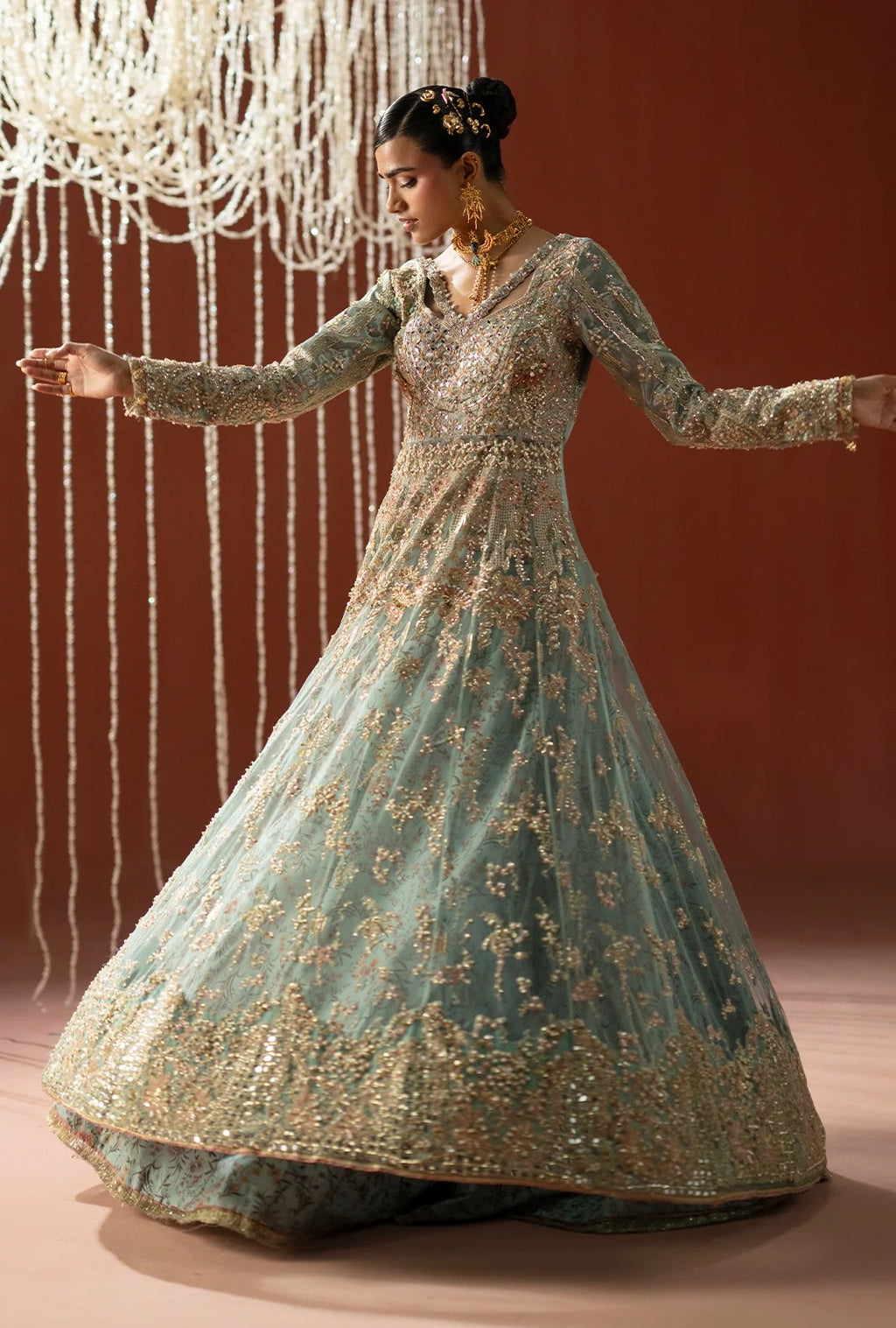A woman modeling a long-sleeved, heavily embroidered light blue-green gown with gold detailing, posed in front of a warm brown backdrop with hanging bead decorations.