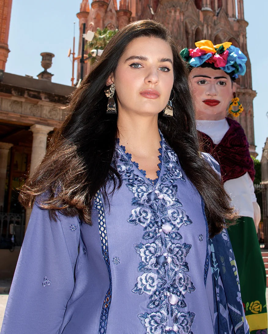 Woman in a blue embroidered outfit standing next to a colorful figurine with a traditional hairstyle, in front of a historic building.