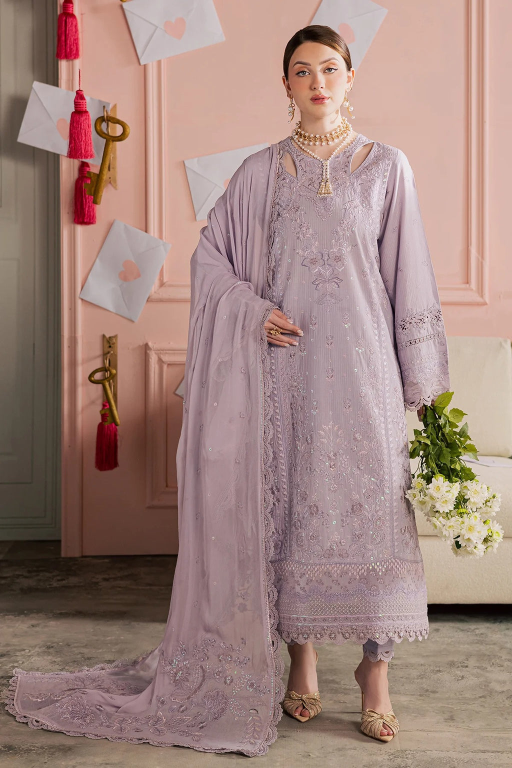 Woman in a lavender embroidered outfit standing in a room with pink walls and decorative elements.