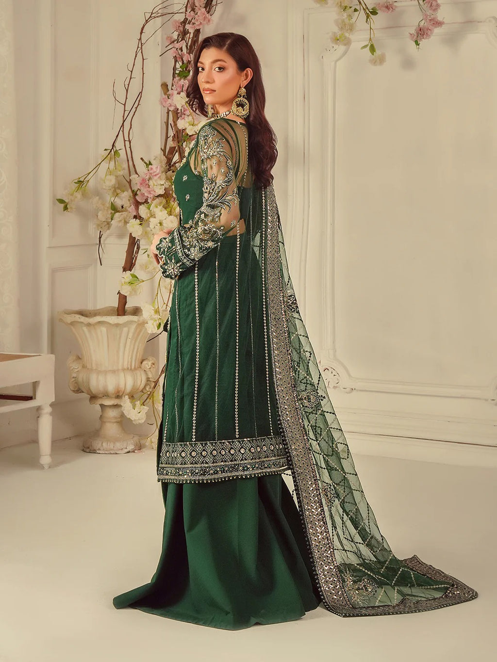Woman in a green traditional outfit with intricate patterns, standing in a decorated room.