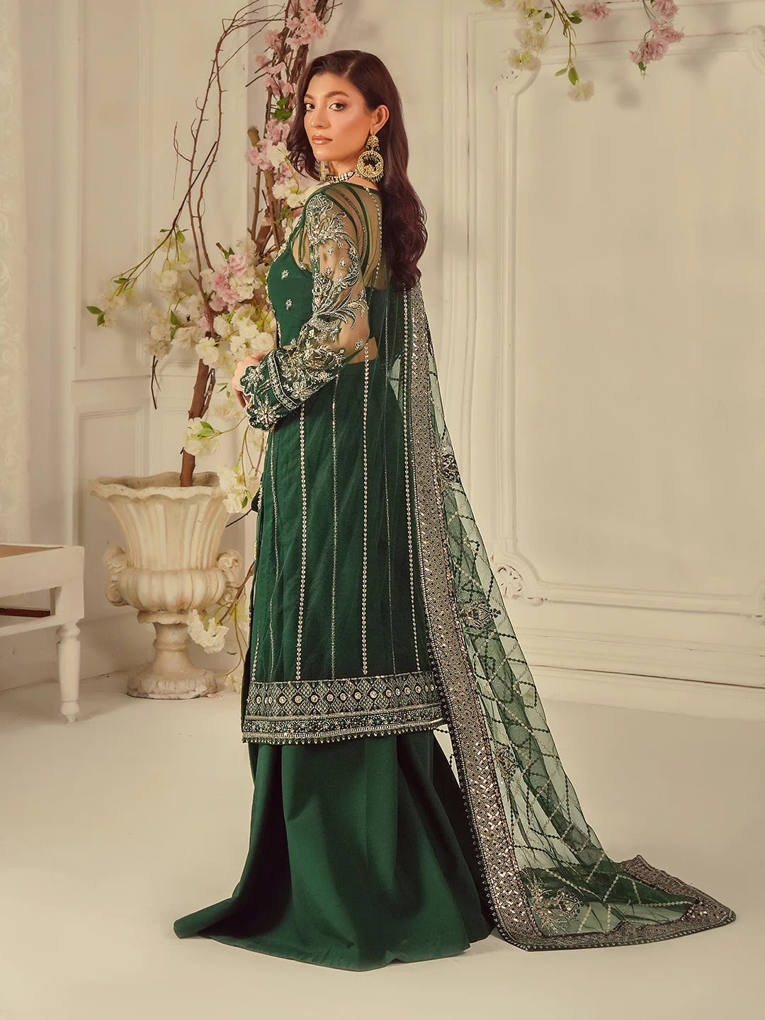 Woman in a green traditional outfit with intricate patterns, standing in a decorated room.