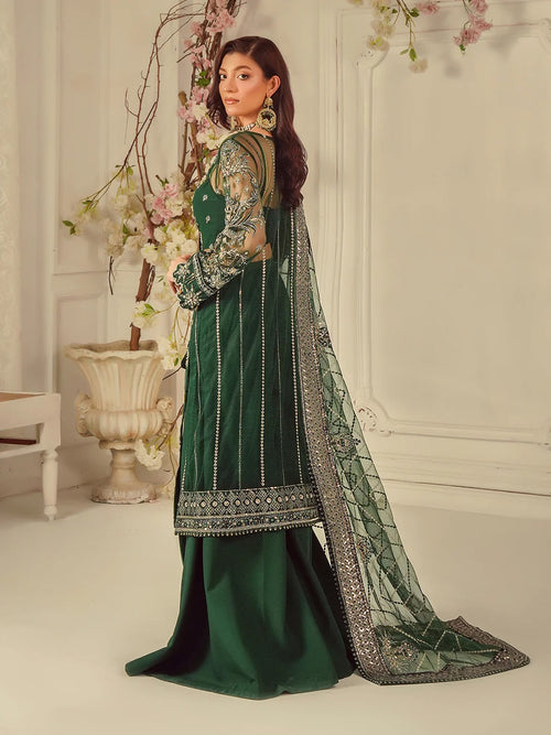Woman in a green traditional outfit with intricate patterns, standing in a decorated room.