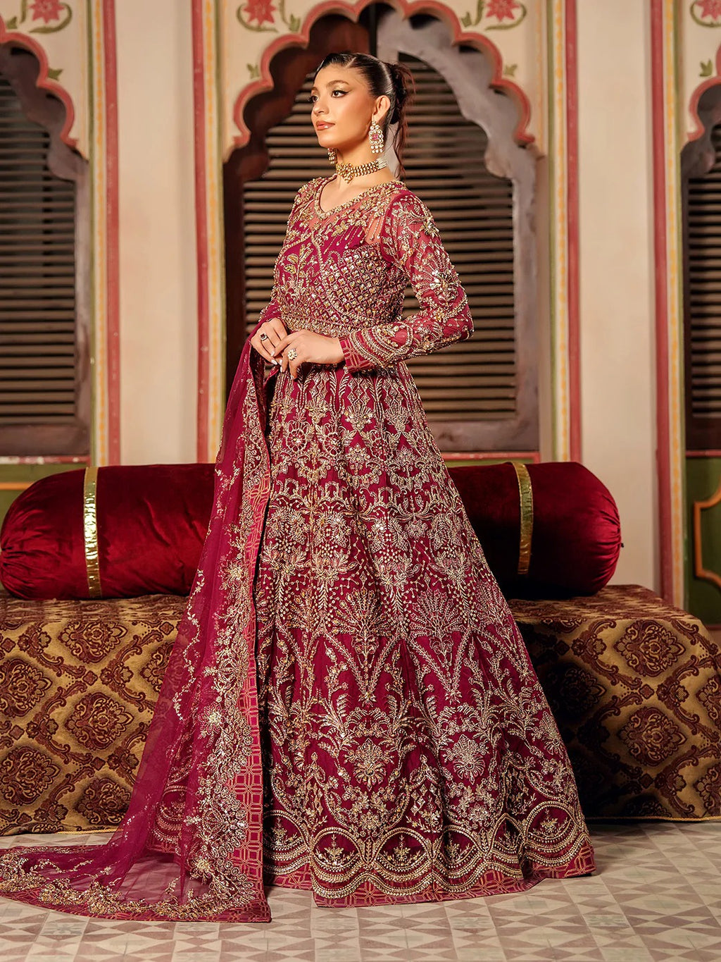 Woman in a traditional maroon and gold embroidered outfit standing in an ornate room.