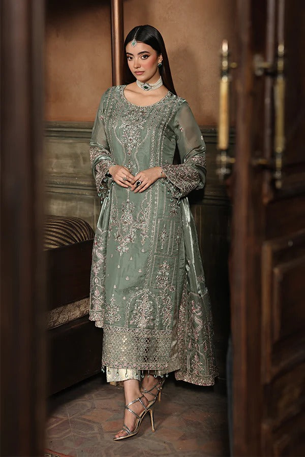 Woman in a green embroidered traditional outfit standing in a room with wooden furniture.