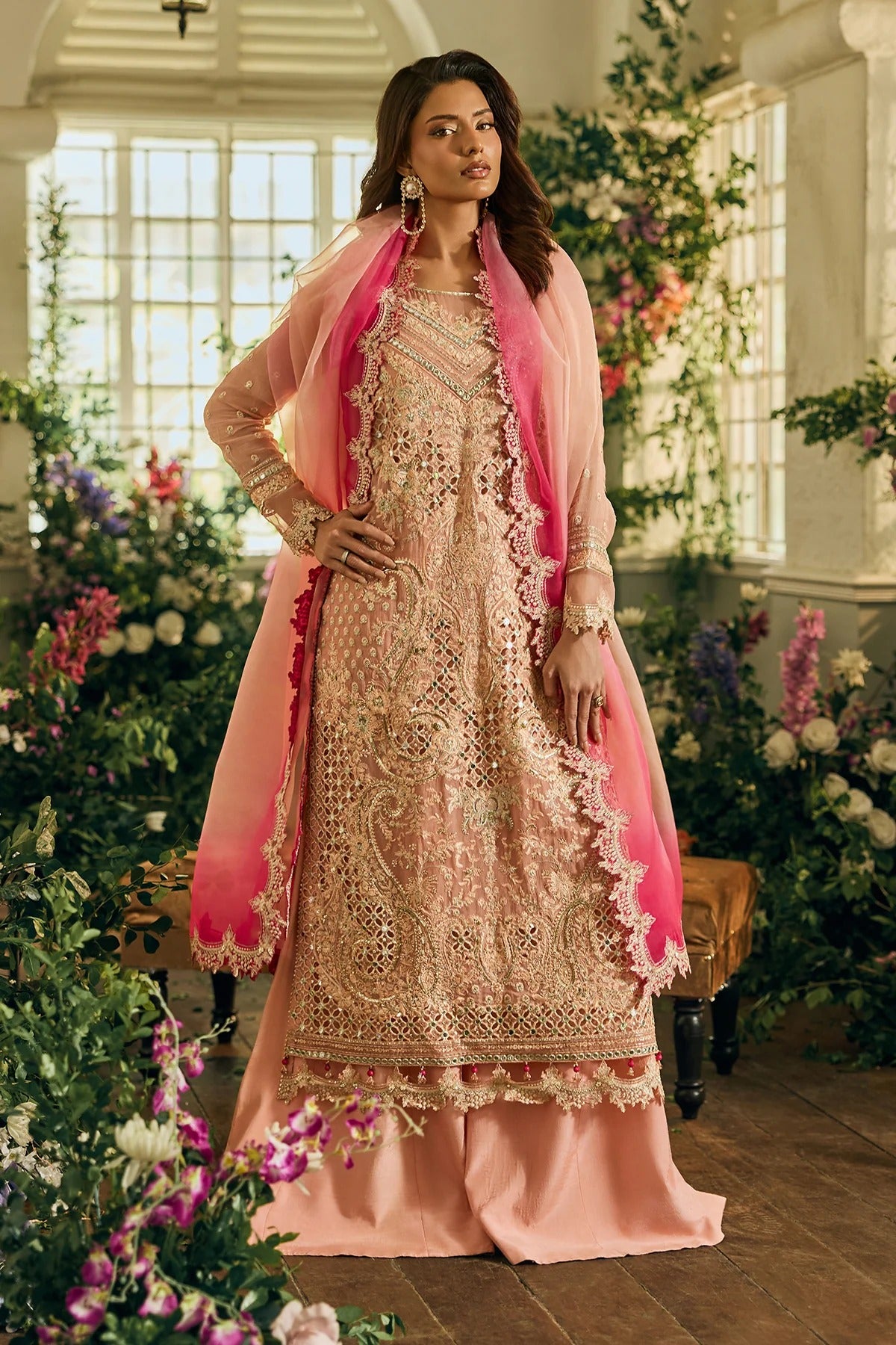 Woman in a traditional outfit with pink dupatta standing in a decorated room with flowers.