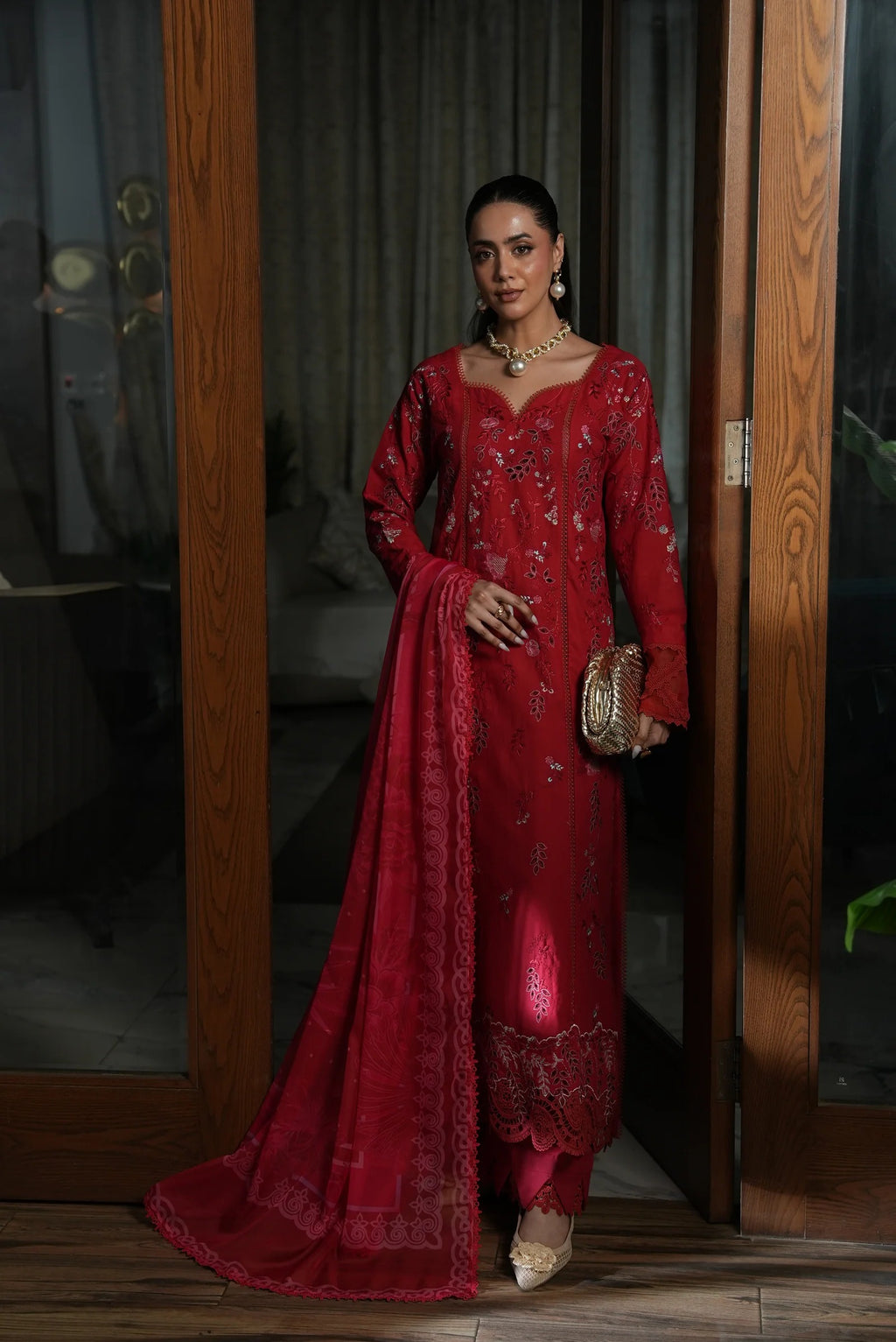 Woman in a red traditional outfit standing indoors with wooden doors and curtains in the background.