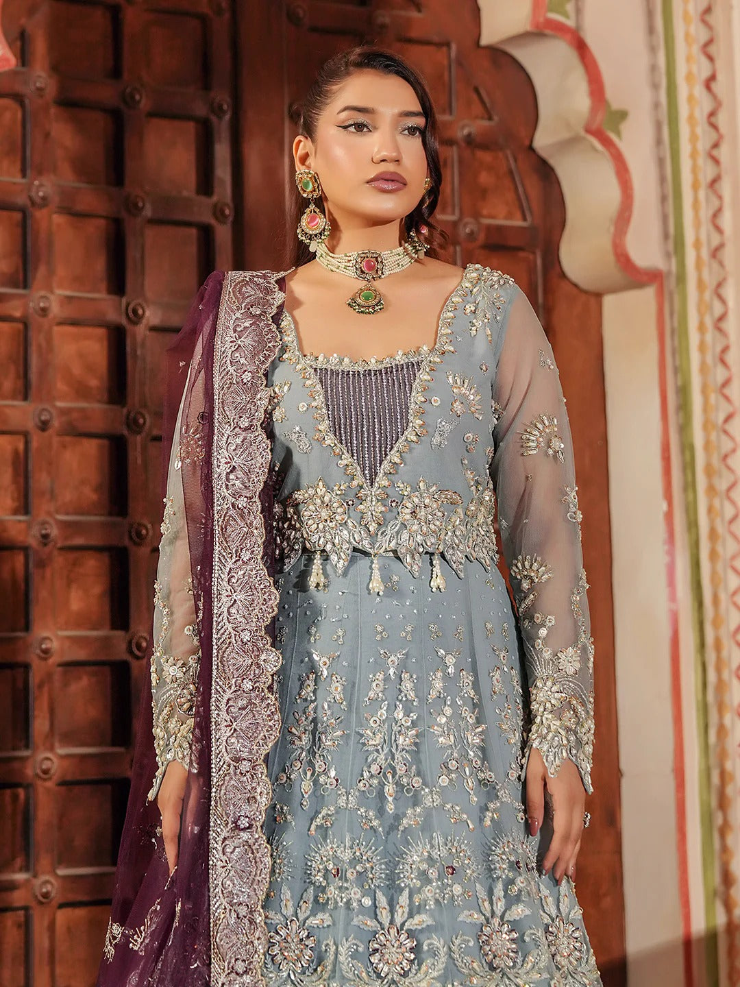 Woman wearing an embroidered traditional outfit with a wooden door and decorative wall in the background