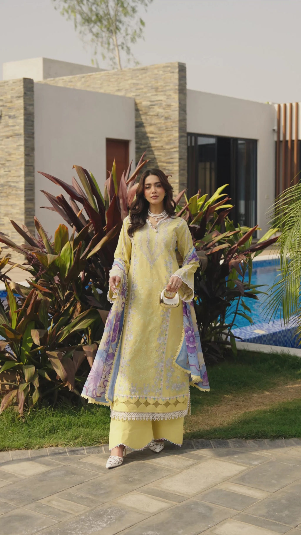 Woman in a light yellow traditional outfit standing outdoors with plants and a building in the background