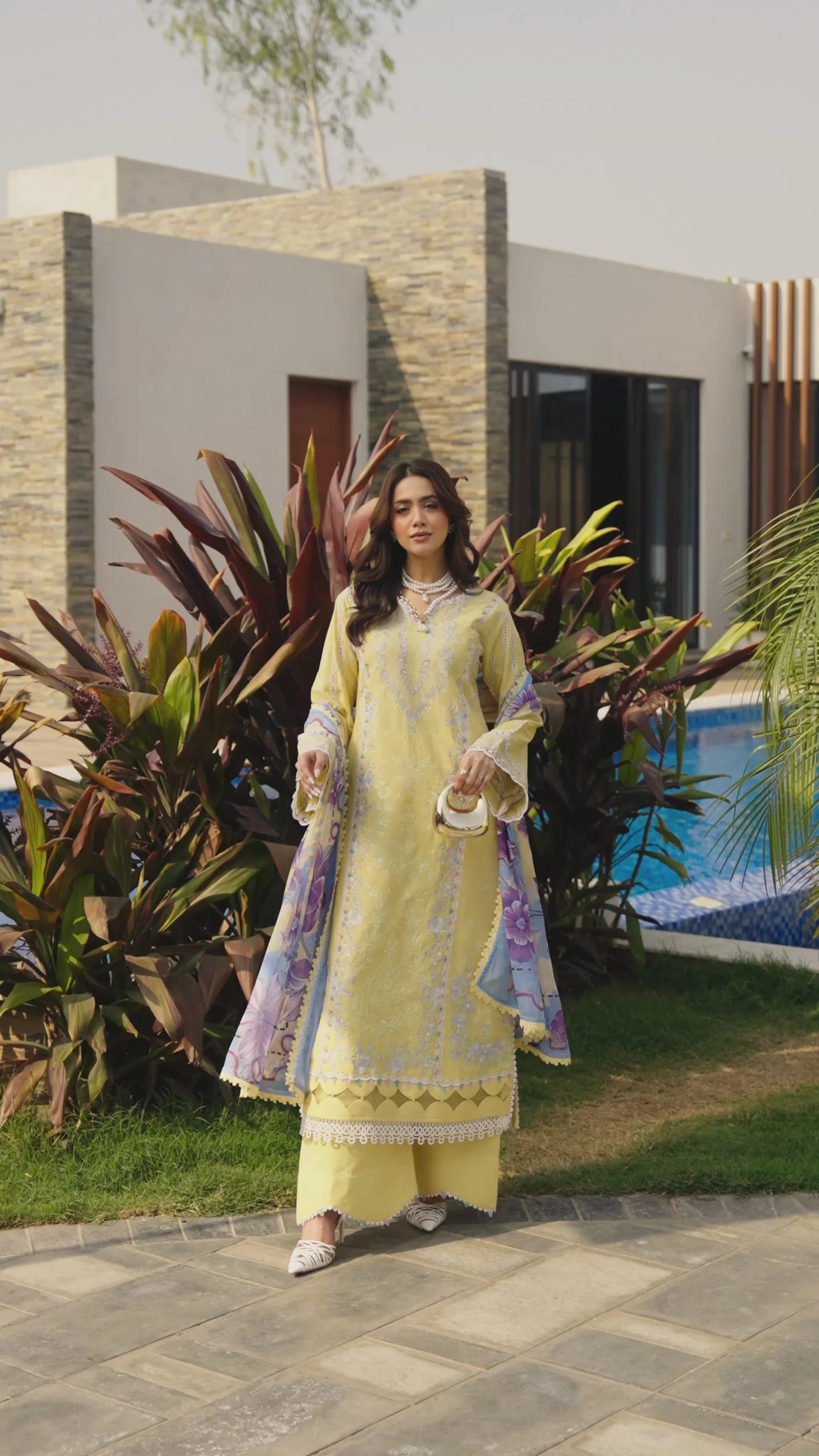 Woman in a light yellow traditional outfit standing outdoors with plants and a building in the background