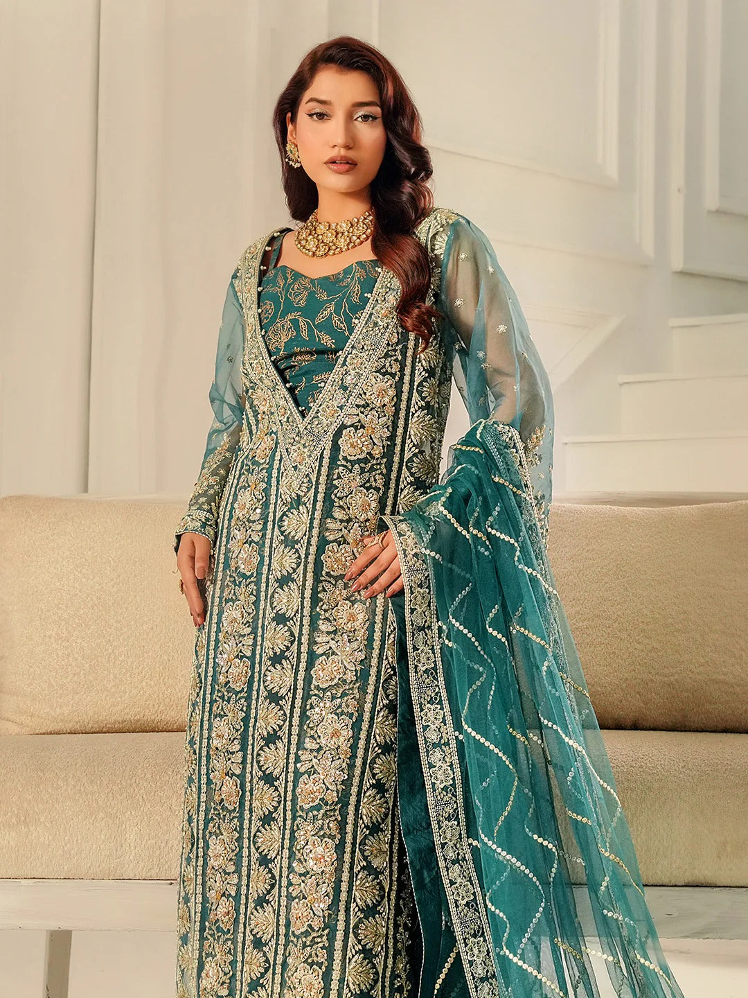 Woman in a teal embroidered traditional outfit with a sheer dupatta in a room.