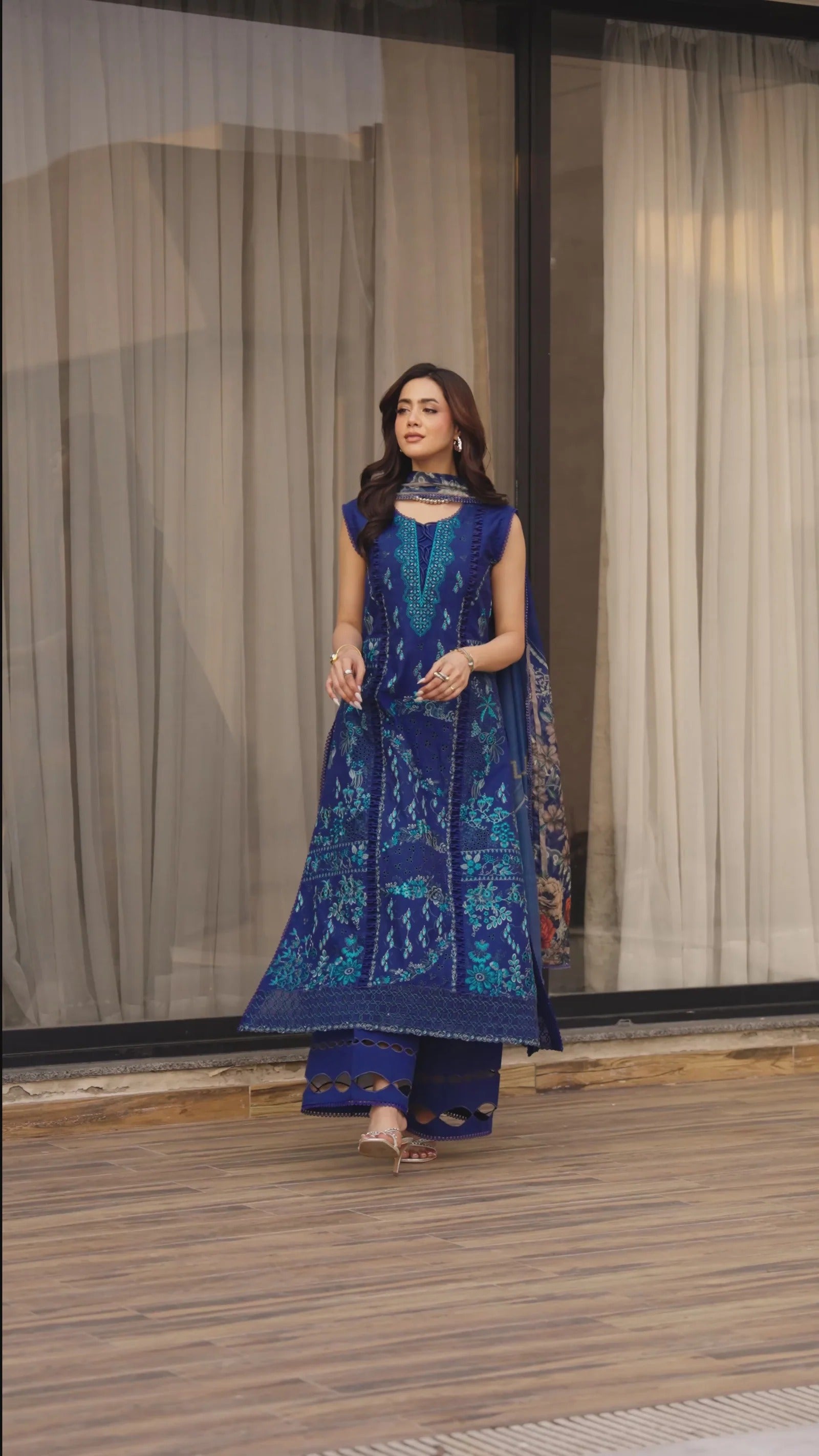 Woman in a blue embroidered outfit walking on a wooden floor with curtains in the background
