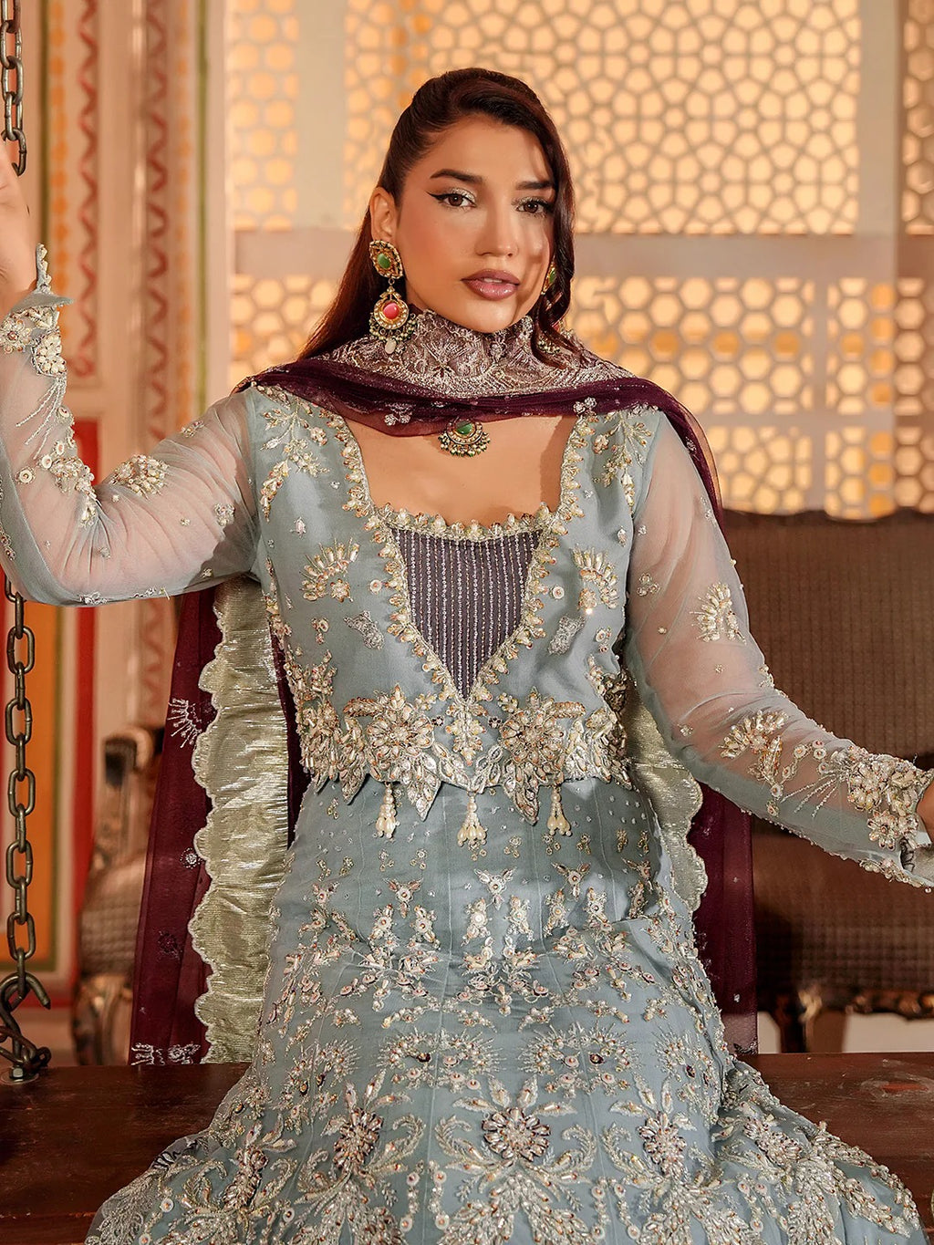 Woman wearing an ornate traditional outfit with intricate designs in a warm-toned interior setting.