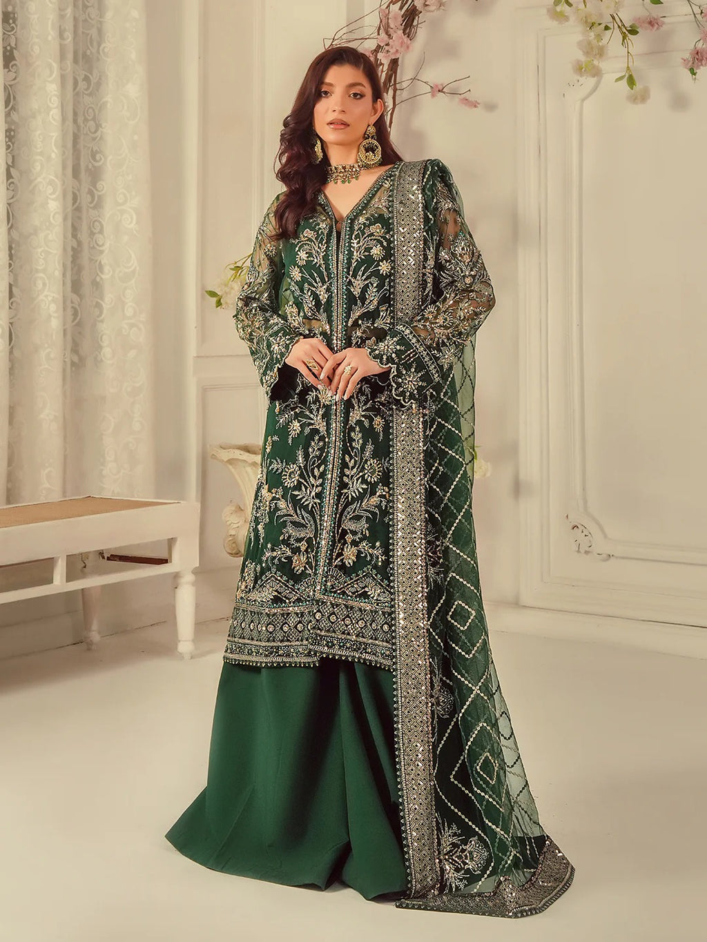 Woman wearing a green and black embroidered traditional outfit in a decorated room.