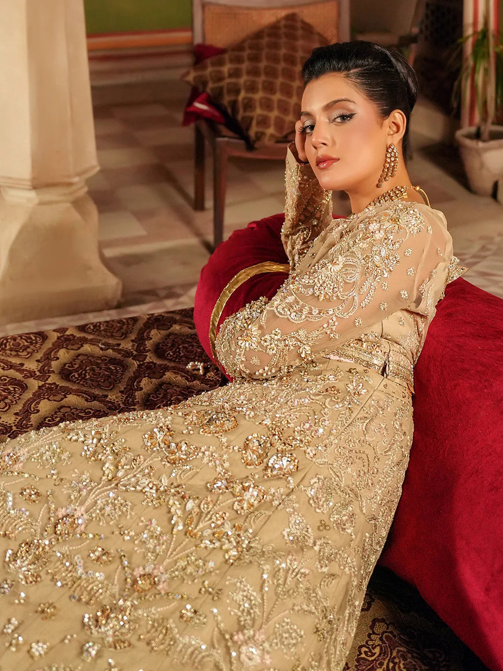 Woman in an ornate gold dress sitting on a red couch in a decorated room.