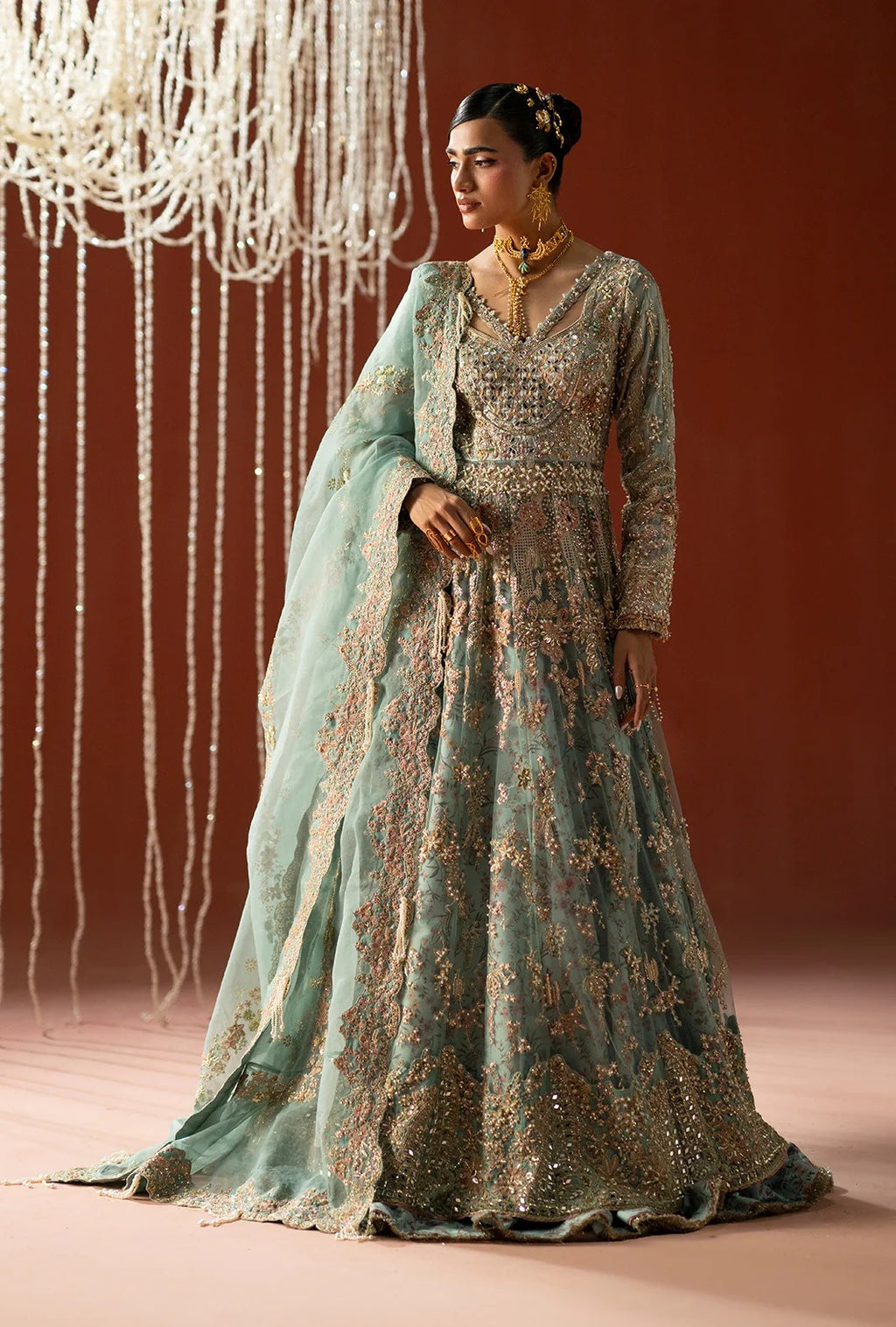 A woman models a traditional long gown with a dusty teal-green base and extensive gold embroidery, paired with a matching sheer dupatta, set against a warm brown backdrop with hanging decorative lights.