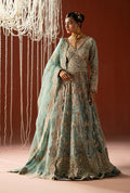 A woman models a traditional long gown with a dusty teal-green base and extensive gold embroidery, paired with a matching sheer dupatta, set against a warm brown backdrop with hanging decorative lights.