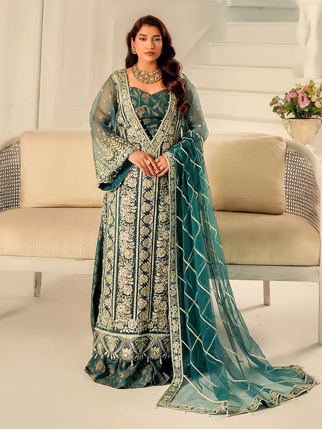 Woman in a teal embroidered traditional outfit standing in a modern interior setting.