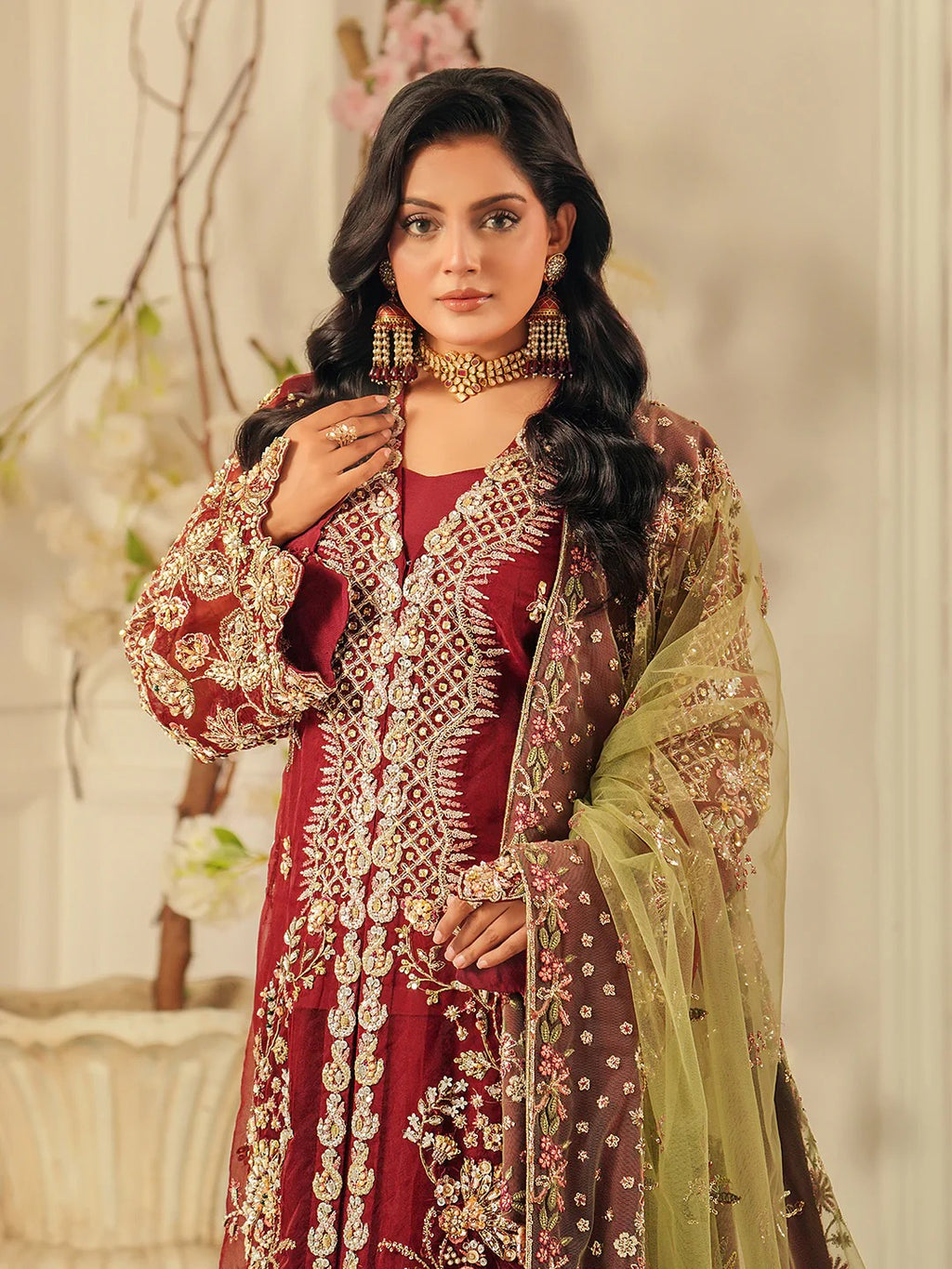Woman in traditional maroon and gold embroidered outfit with green dupatta against a neutral background